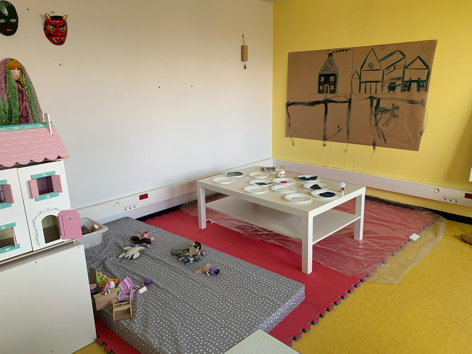 Salle d'art-thérapie pour enfants à l'IDEFHI avec maison de poupée, masques au mur, table basse et tapis coloré