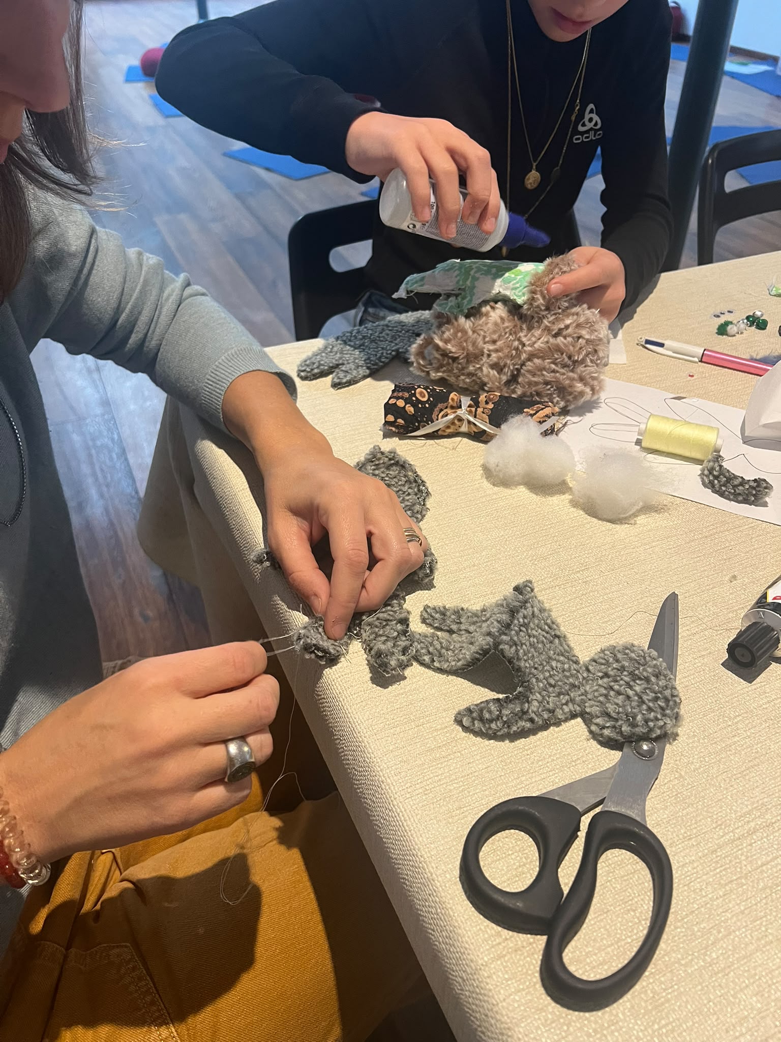 Mains cousant de petites marionnettes en tissu gris et brun sur une table d'atelier, avec ciseaux et fil