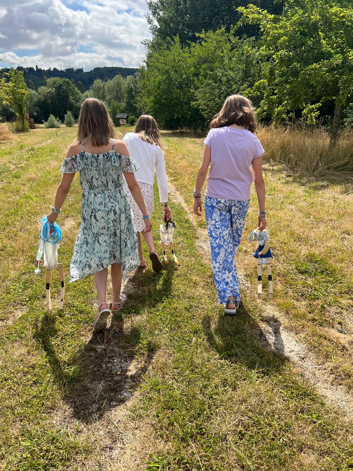 Trois personnes vues de dos marchant dans un pré ensoleillé, portant chacune une marionnette artisanale
