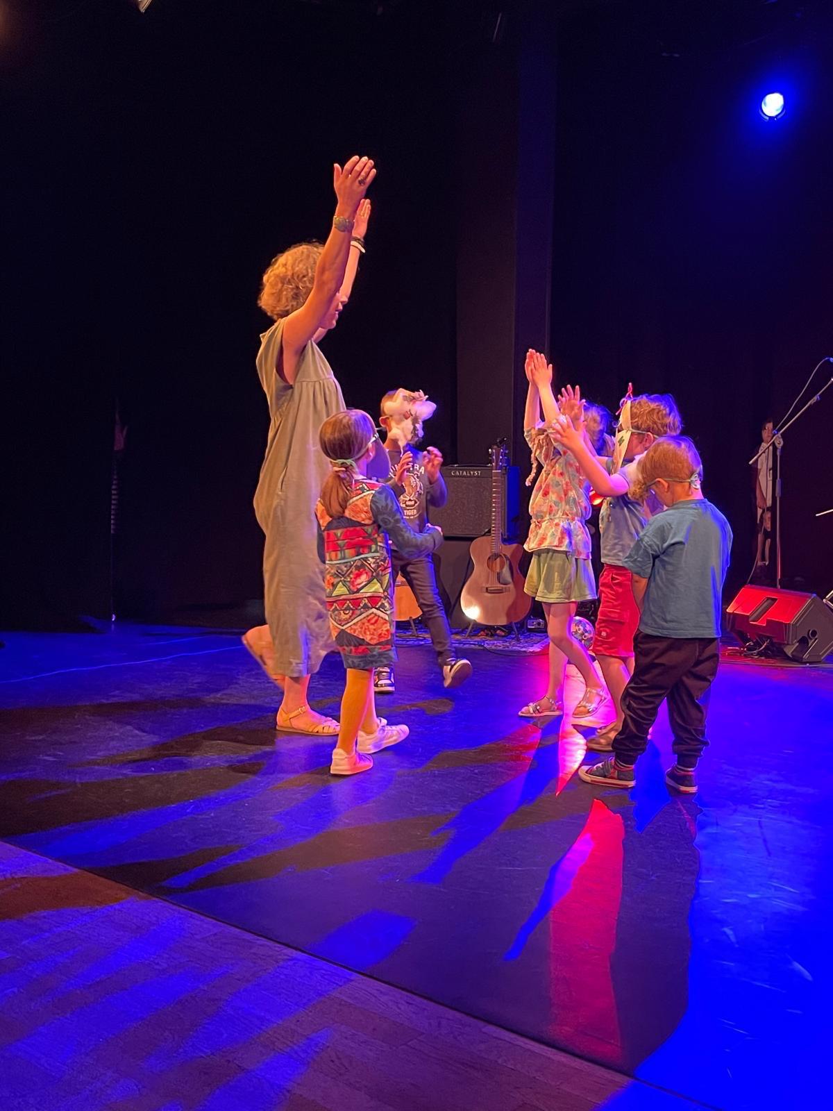 Raphaëlle et un groupe de jeunes enfants dansant et levant les mains sur la scène d'une salle de spectacle, guitare et éclairages bleus en fond