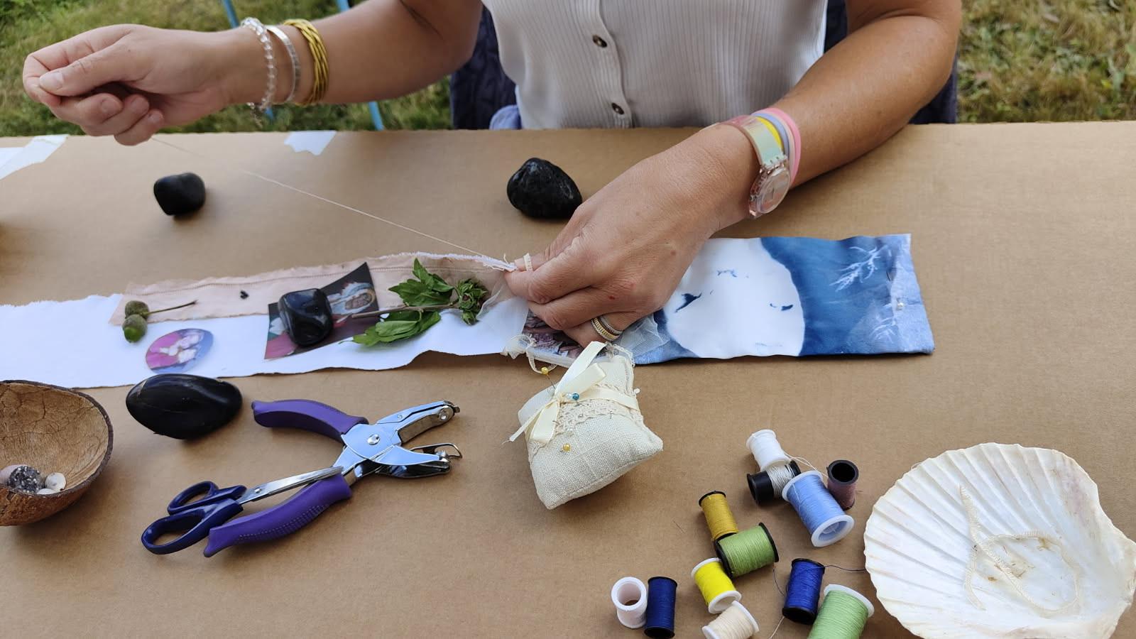 Mains assemblant tissus, fils colorés, pierres et coquillage sur une table en plein air
