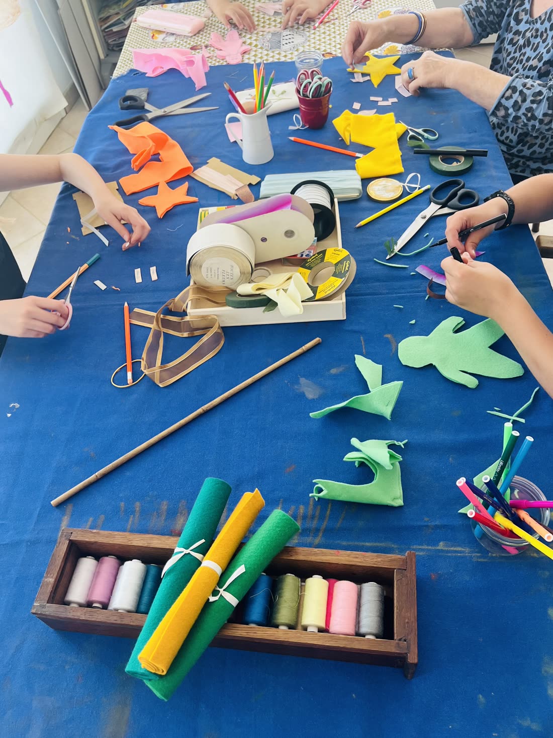Table d'atelier couverte de feutrine colorée, ciseaux, rubans et bobines de fil, plusieurs mains au travail