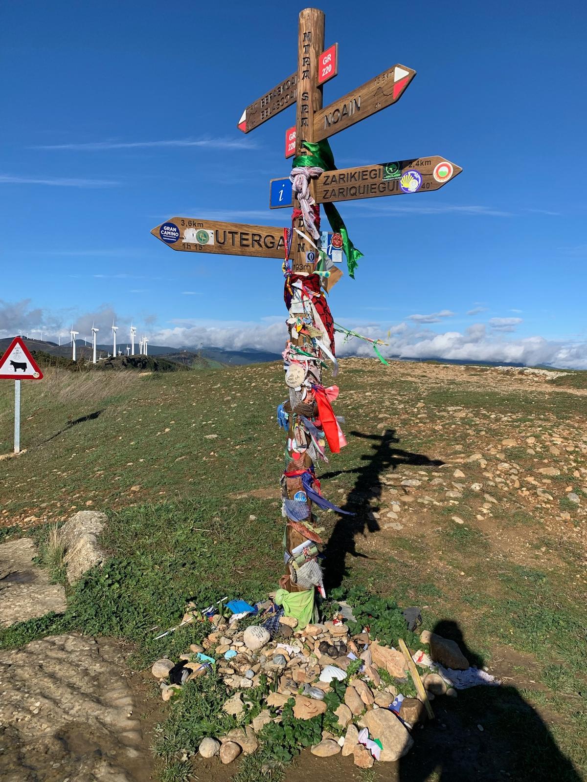 Poteau directionnel de randonnée couvert de rubans et souvenirs, sur un chemin en pleine nature sous un ciel bleu